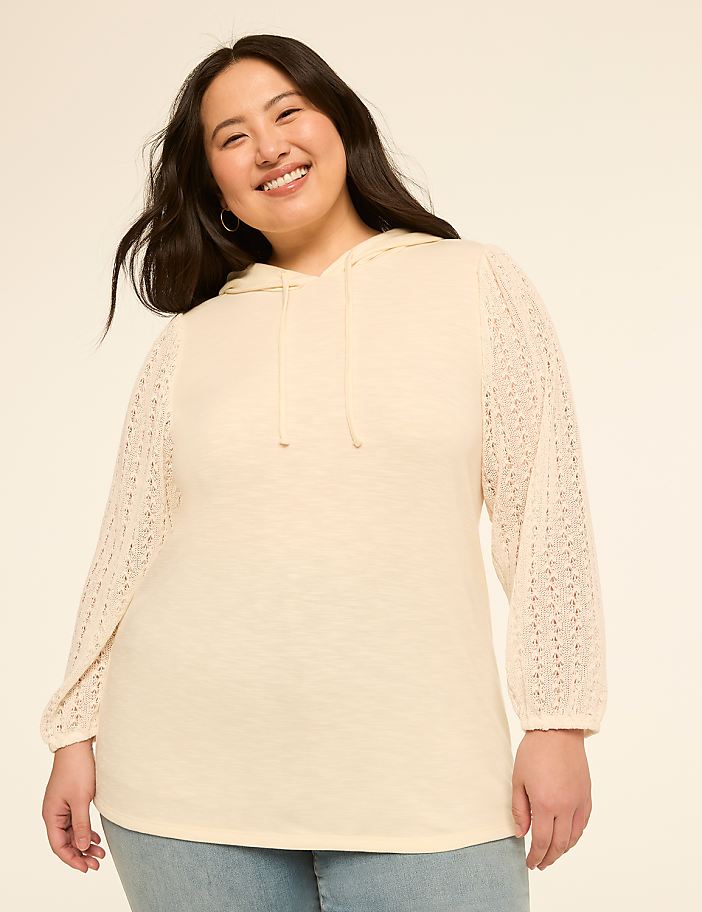 A smiling woman in a cream-colored hoodie with lace sleeves stands against a plain, light background.
