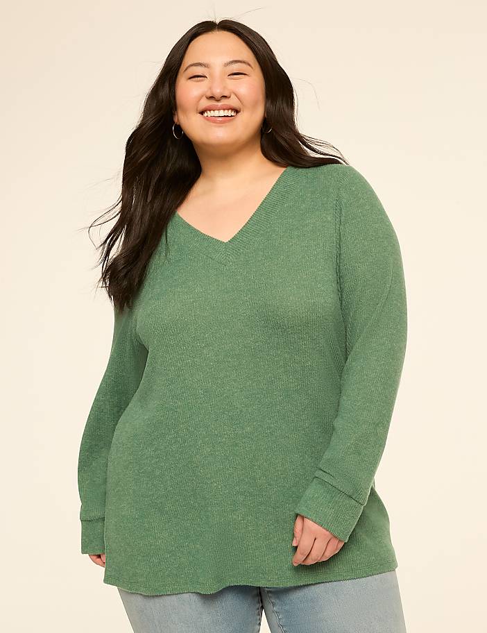 A smiling woman in a green v-neck sweater stands against a plain, light-colored background.