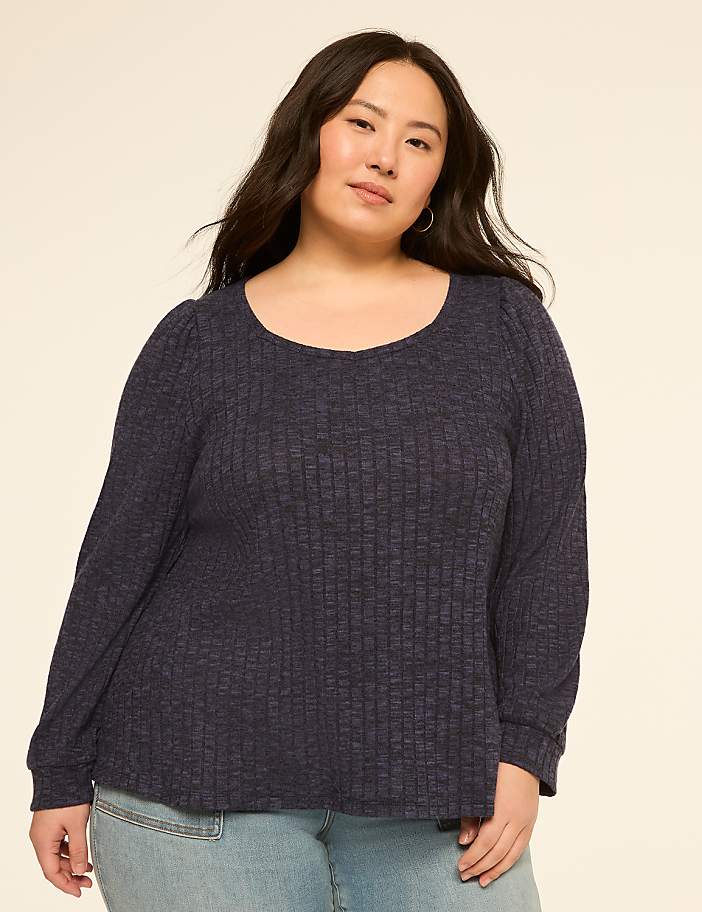 A woman with dark hair and a blue ribbed sweater stands against a plain, light background.