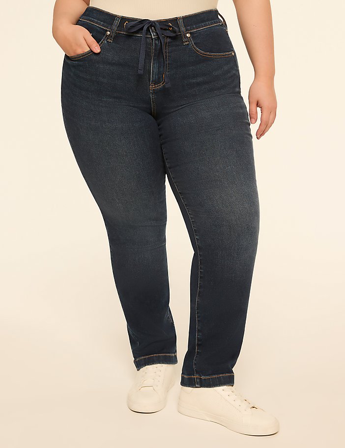 A person in dark wash jeans and white sneakers stands against a plain, light-colored background.