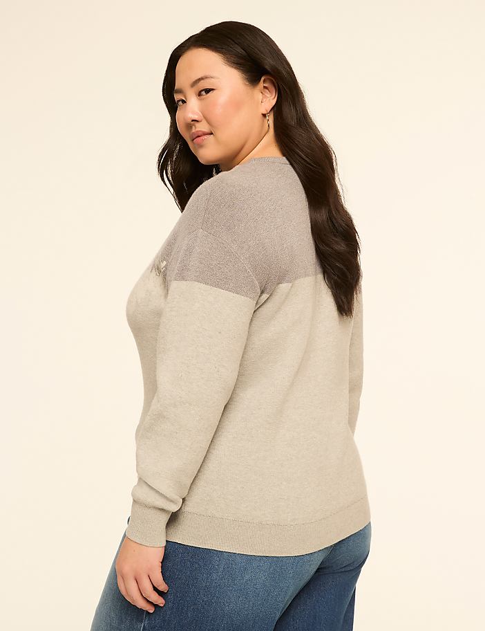 A woman in a two-toned sweater and jeans poses against a plain, light-colored background.