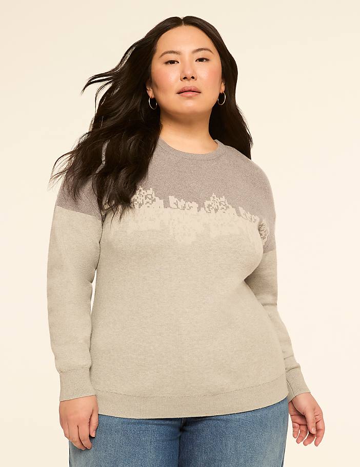 A woman with dark hair and hoop earrings stands in the foreground wearing a two-toned gray sweater with a lace-like pattern across the chest, against a plain, light beige background.