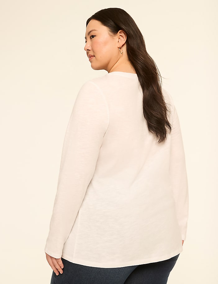 A woman in a white long-sleeved shirt and dark jeans stands against a plain, light-colored background.