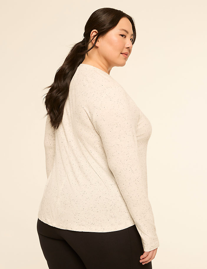 A woman with dark hair pulled back in a ponytail, wearing a speckled cream-colored long-sleeved shirt and black pants, is shown against a plain, light-colored background.