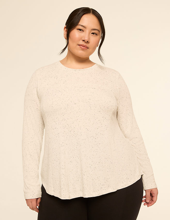 A woman with dark hair pulled back wears a speckled cream long-sleeved shirt and black leggings against a plain, light-colored background.