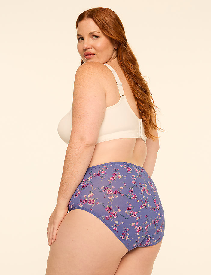 A woman with red hair poses in a cream-colored bra and floral-patterned underwear against a plain, light background.