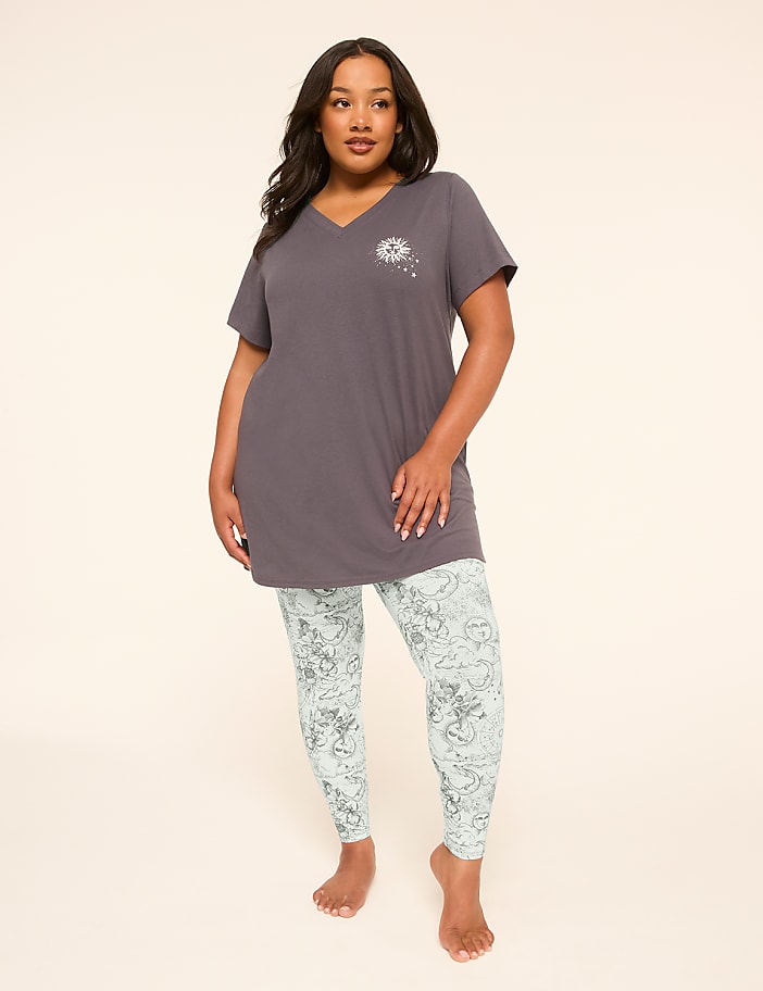 A woman stands in the foreground wearing a grey tunic and patterned leggings against a plain, light-colored background.