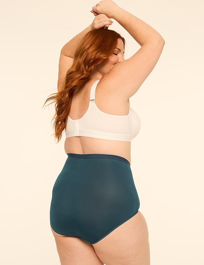 A woman with red hair in a white bra and teal underwear poses with her arms raised against a plain, light-colored background.