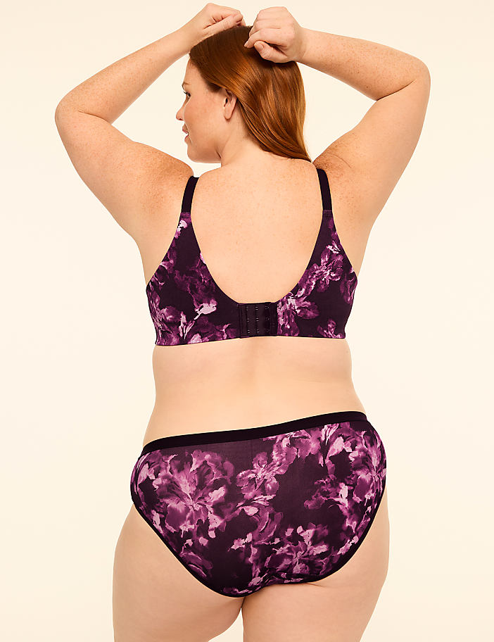 A woman with red hair, seen from the back, wears a floral purple bra and matching underwear against a plain, light-colored background.