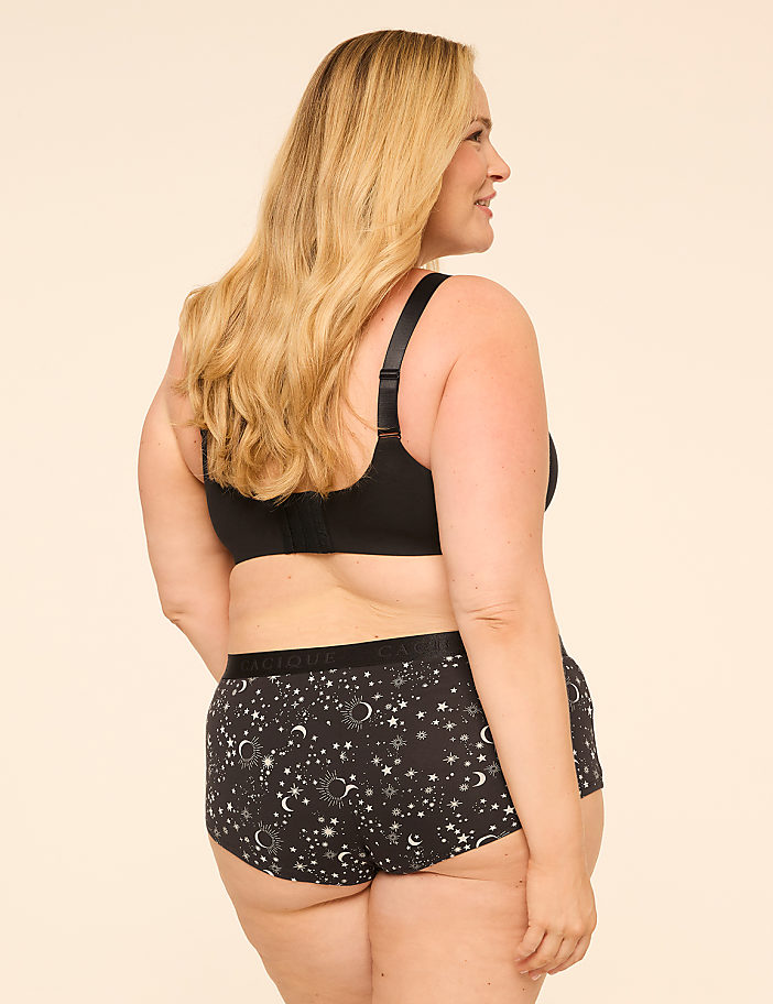 A woman in a black bra and patterned underwear stands against a plain, light-colored background.