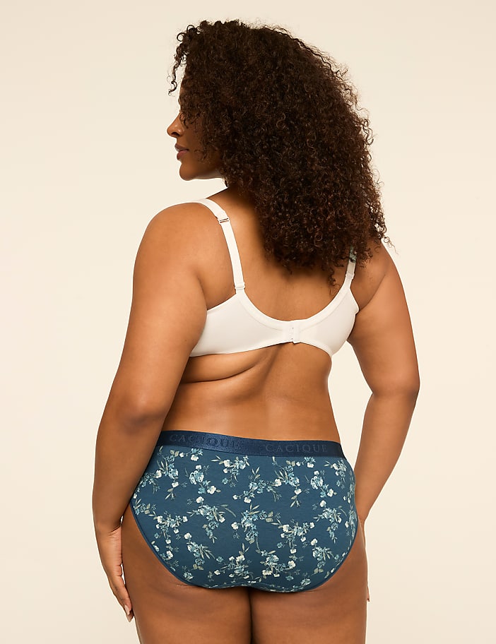 A woman in a white bra and floral-patterned underwear stands against a plain, light-colored background.