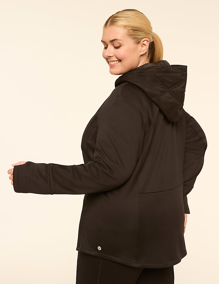 A woman in a black hooded jacket smiles over her shoulder against a plain, light-colored background.
