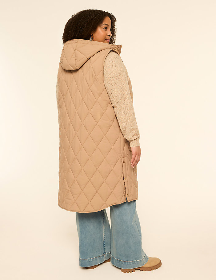 A woman in a tan quilted vest and light blue wide-leg jeans stands against a plain cream background.