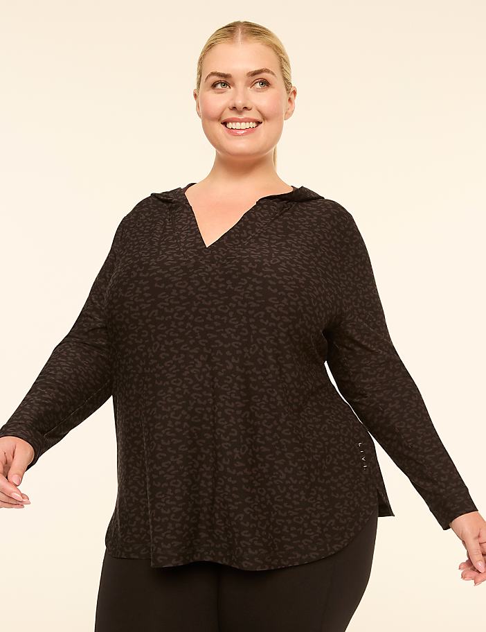 A smiling woman in a dark, leopard-print hooded top stands against a plain, light-colored background.