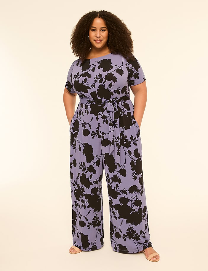 A woman with curly hair stands in a floral jumpsuit with her hands in her pockets against a plain, light-colored background.