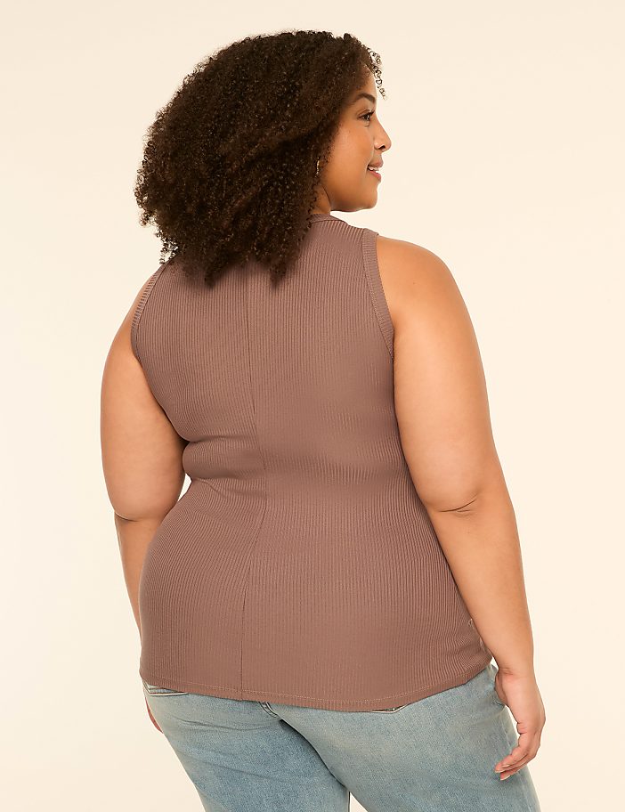 A woman with curly brown hair wearing a brown ribbed tank top and blue jeans stands against a plain cream background.