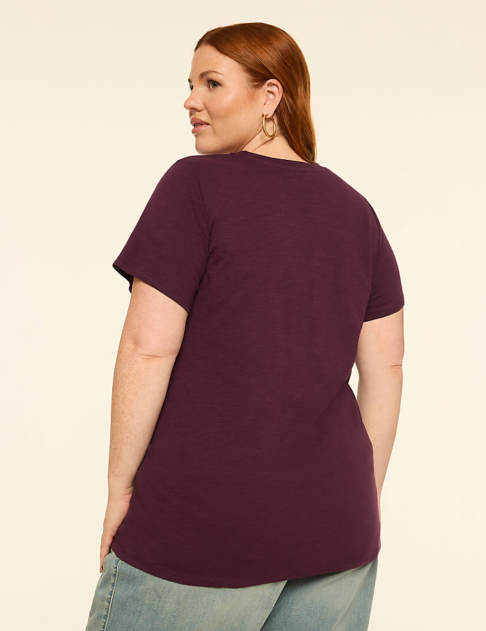 A woman with red hair wearing a purple t-shirt and jeans stands against a plain, light-colored background.