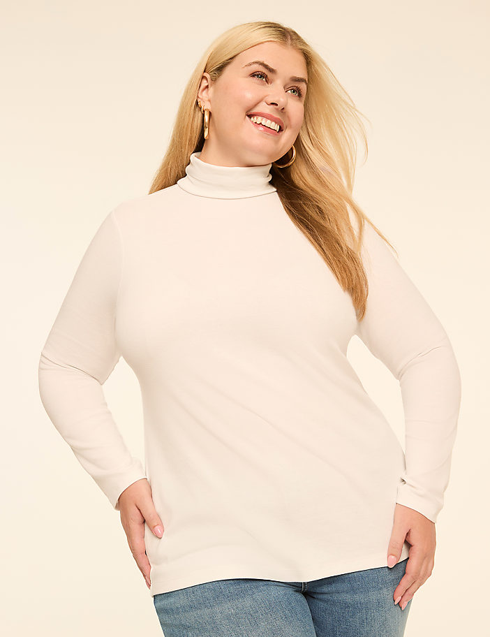 A smiling woman with blonde hair wears a white turtleneck and blue jeans, posing against a plain, light-colored background.
