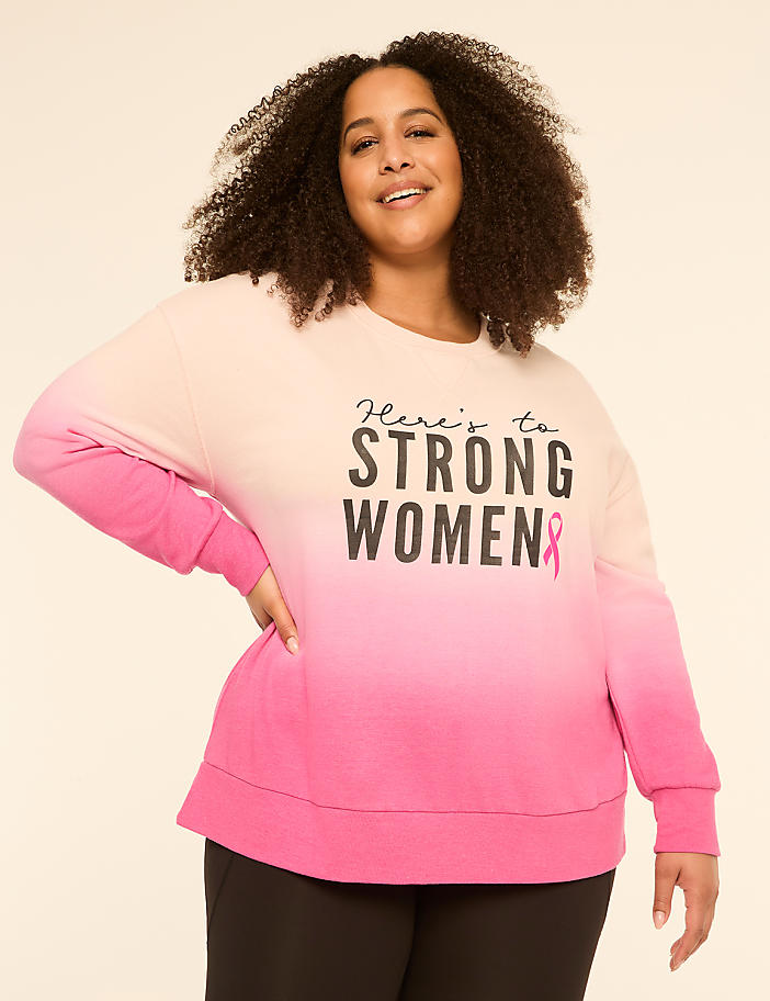 A smiling woman with curly hair poses in a pink and cream ombre sweatshirt with the words "Here's to STRONG WOMEN" and a pink ribbon, against a plain, light-colored background.