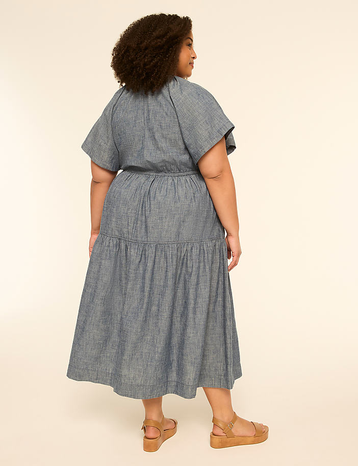 A woman in a blue tiered dress and sandals stands against a plain, light-colored background.