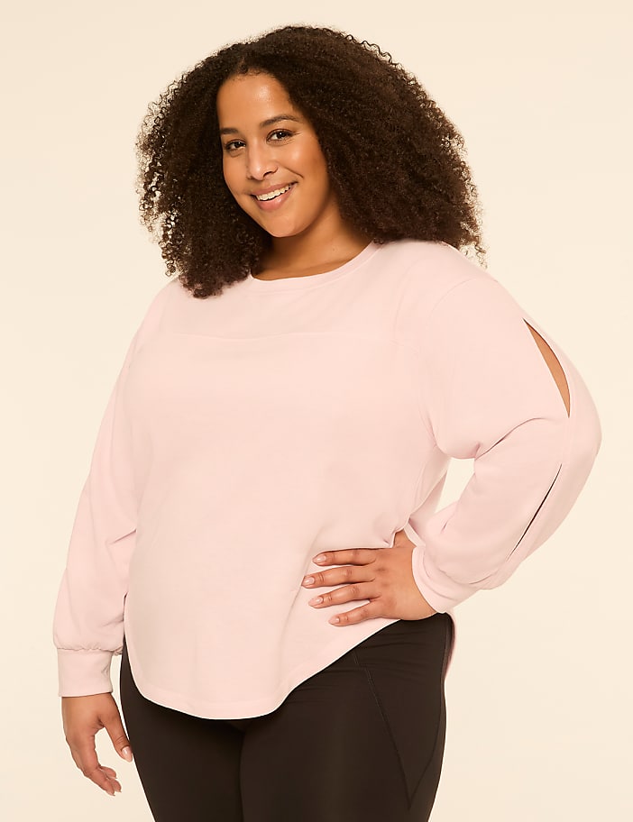 A smiling woman with curly hair poses in a light pink top and black leggings against a plain, light background.