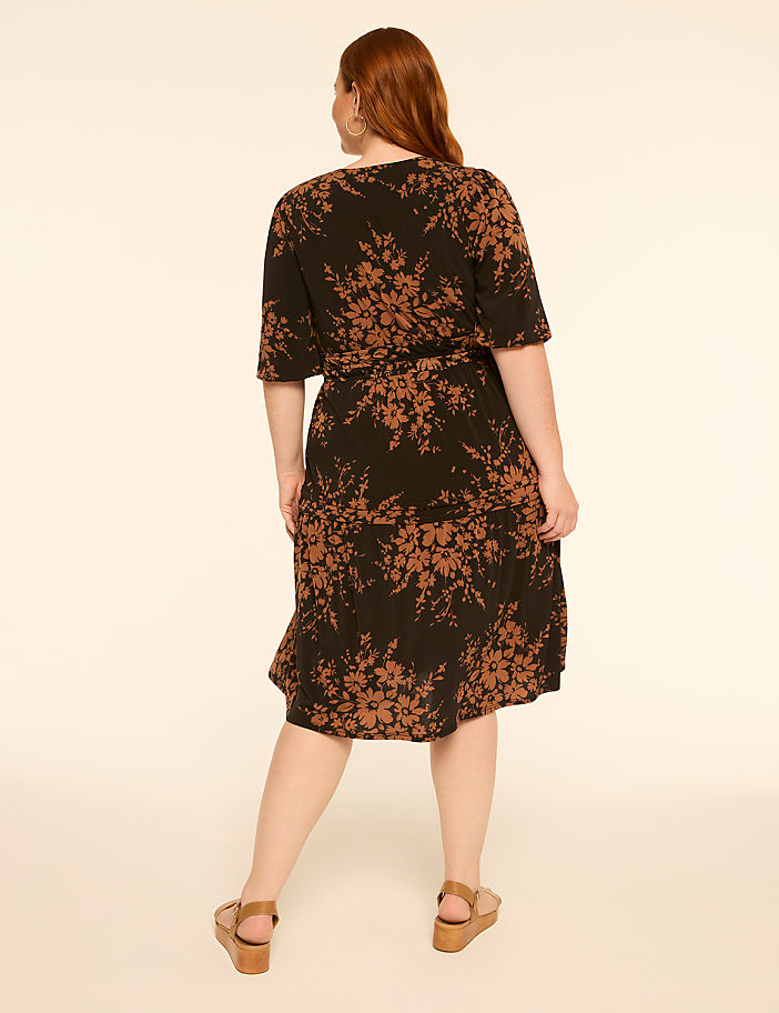 A woman in a floral print dress stands against a plain, light-colored background.