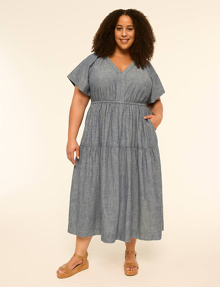 A smiling woman in a blue tiered dress stands against a plain, light-colored background.