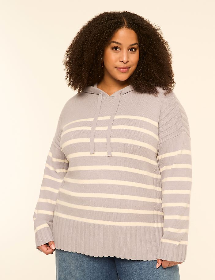 A woman with curly hair wears a striped hoodie and jeans against a plain, light background.