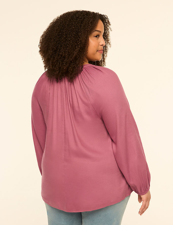 A woman with curly brown hair, seen from the back, wears a mauve peasant blouse and light blue jeans against a plain cream background.
