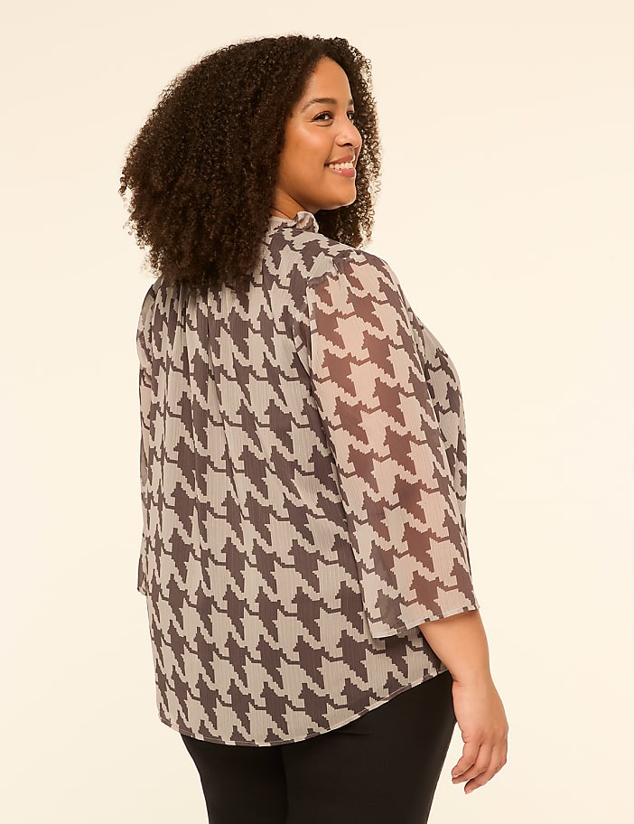 A woman with curly dark hair smiles over her shoulder, wearing a sheer houndstooth blouse and black pants against a plain, light-colored background.