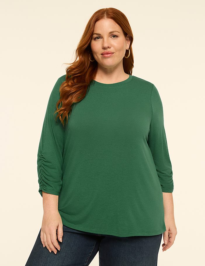 A woman with auburn hair and a green top stands against a plain, light-colored background.
