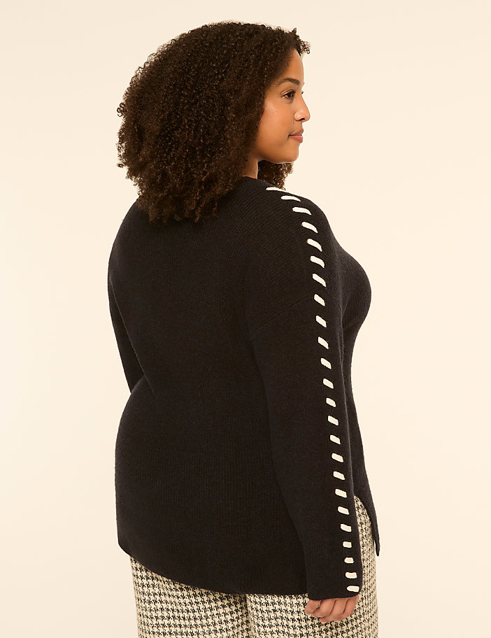 A woman in a dark sweater with white stitching on the sleeves stands against a plain, light-colored background.