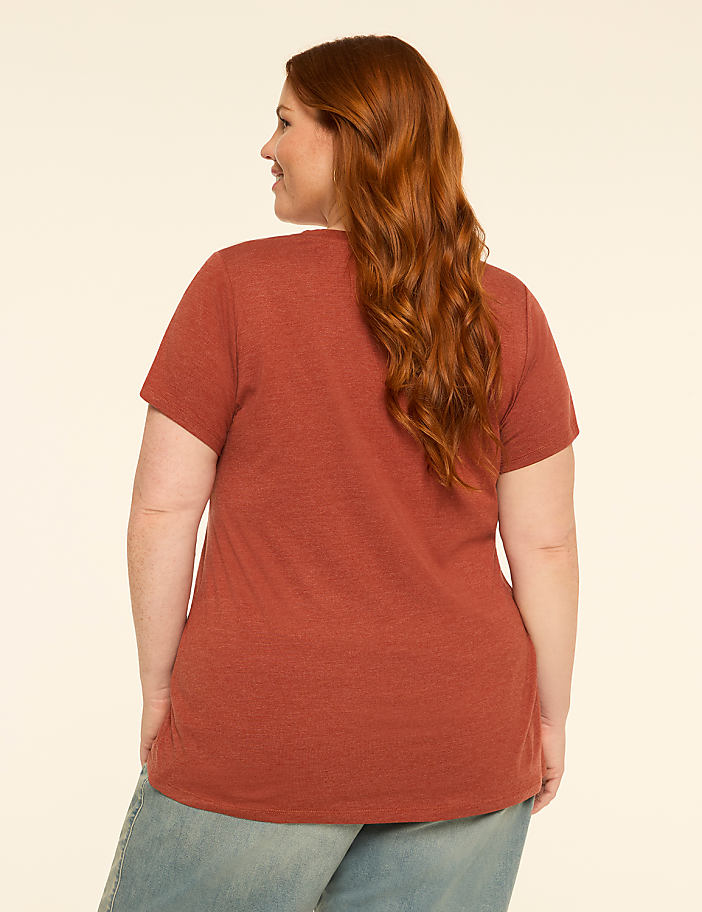 A woman with auburn hair, wearing a rust-colored t-shirt and light blue jeans, stands against a plain, light-colored background.