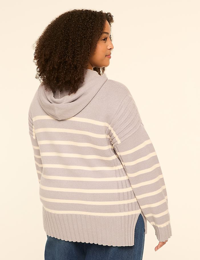 A young woman with curly hair, wearing a striped hooded sweater and blue jeans, stands against a plain, light-colored background.