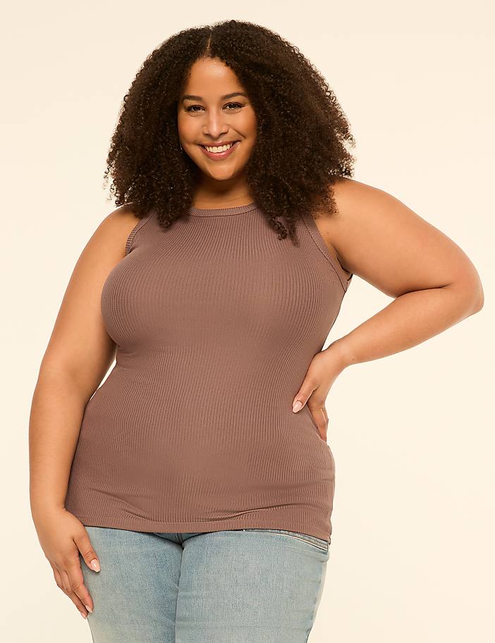 A smiling woman with curly hair in a brown tank top and jeans stands against a plain, light-colored background.