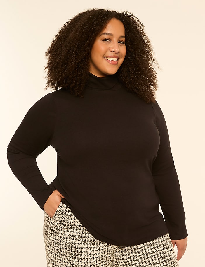 A smiling woman with curly hair in a black turtleneck and patterned pants stands against a plain, light-colored background.
