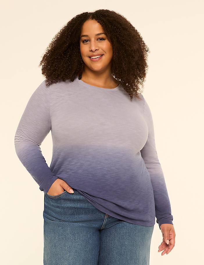 A smiling woman with curly hair in a blue ombre long-sleeved shirt and jeans stands against a plain cream background.