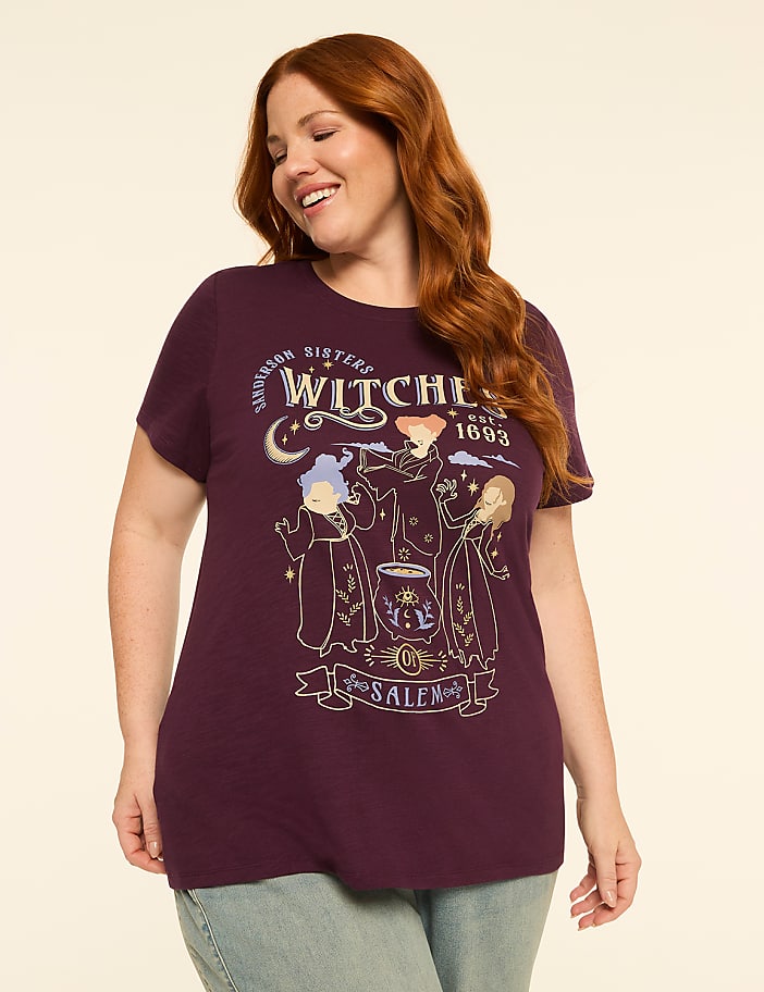 A smiling woman with auburn hair wears a plum t-shirt with a graphic of three witches and a cauldron, set against a plain cream background.