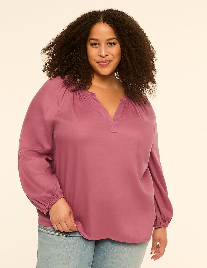 A woman with curly hair smiles at the camera, wearing a mauve blouse and jeans against a plain, light background.