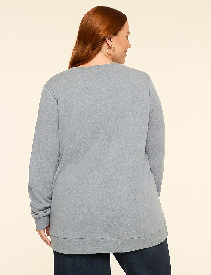 A woman with red hair wearing a gray sweatshirt and dark jeans stands against a plain, light-colored background.