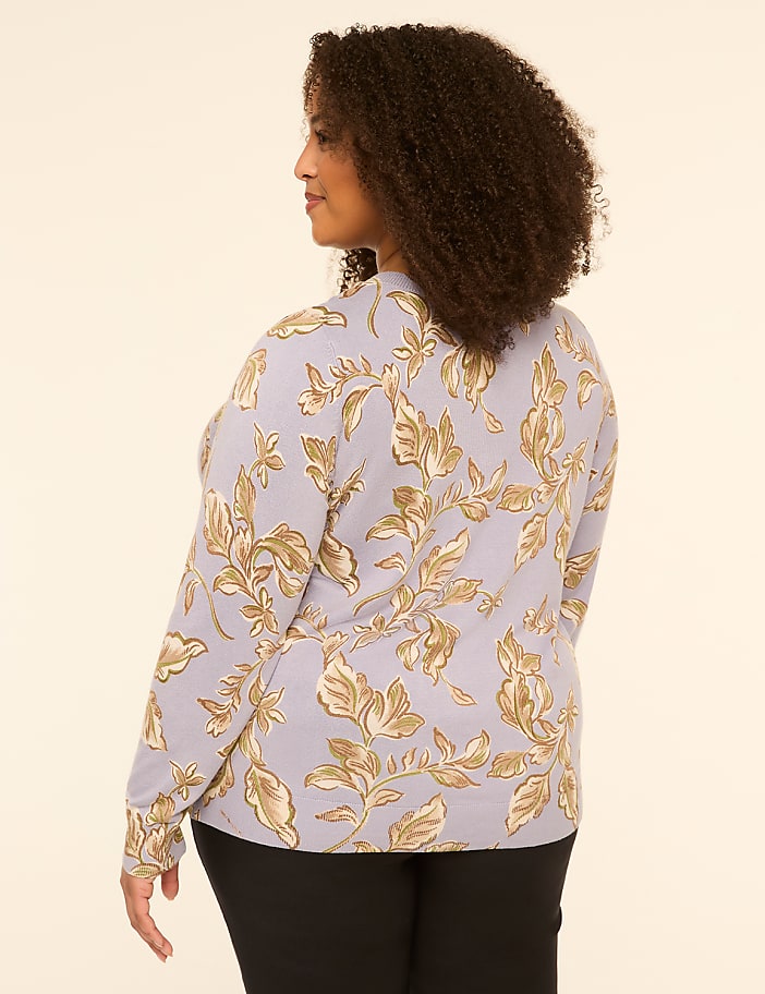 A woman with curly brown hair, wearing a light purple floral-patterned sweater and black pants, stands against a plain beige background.