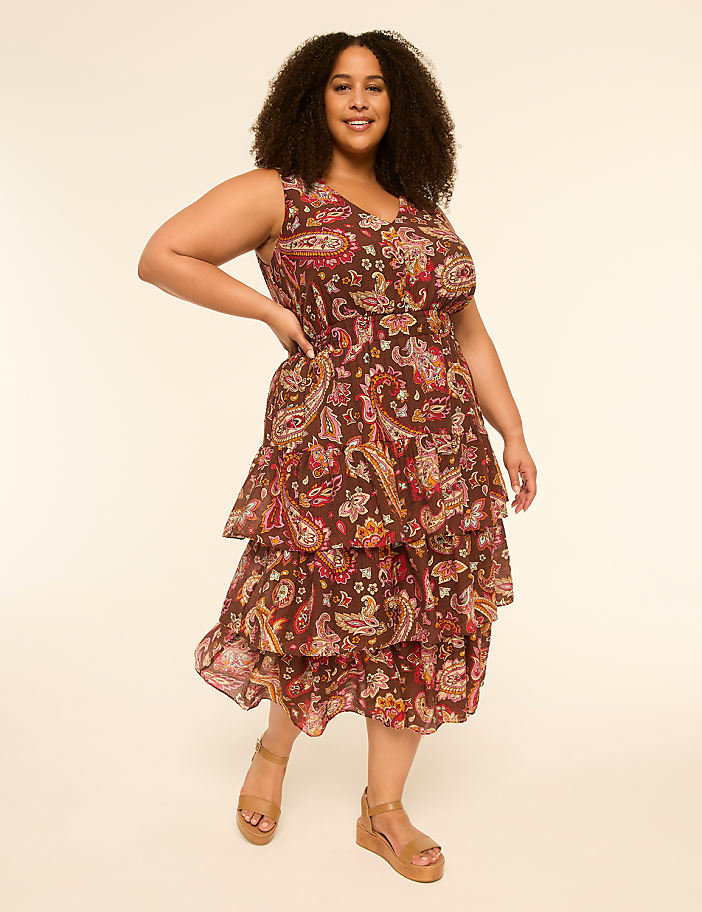 A woman in a tiered paisley dress and wedge sandals stands against a plain, light-colored background.