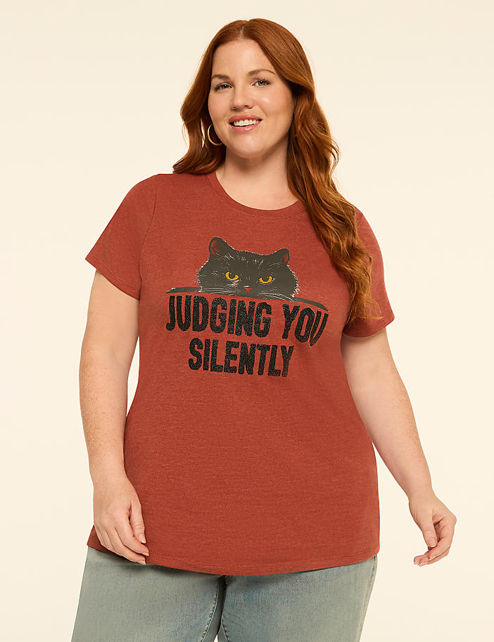 A woman with auburn hair smiles at the camera, wearing a rust-colored t-shirt with a black cat graphic that reads "JUDGING YOU SILENTLY," against a plain, light-colored background.