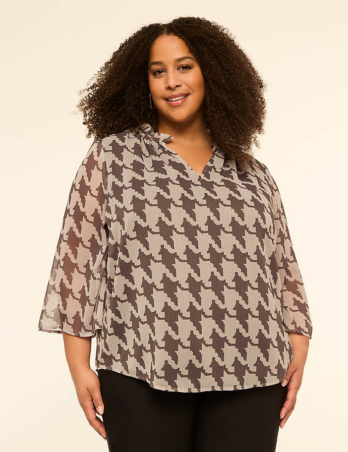 A woman with curly hair smiles at the camera, wearing a patterned blouse and black pants against a plain, light-colored background.