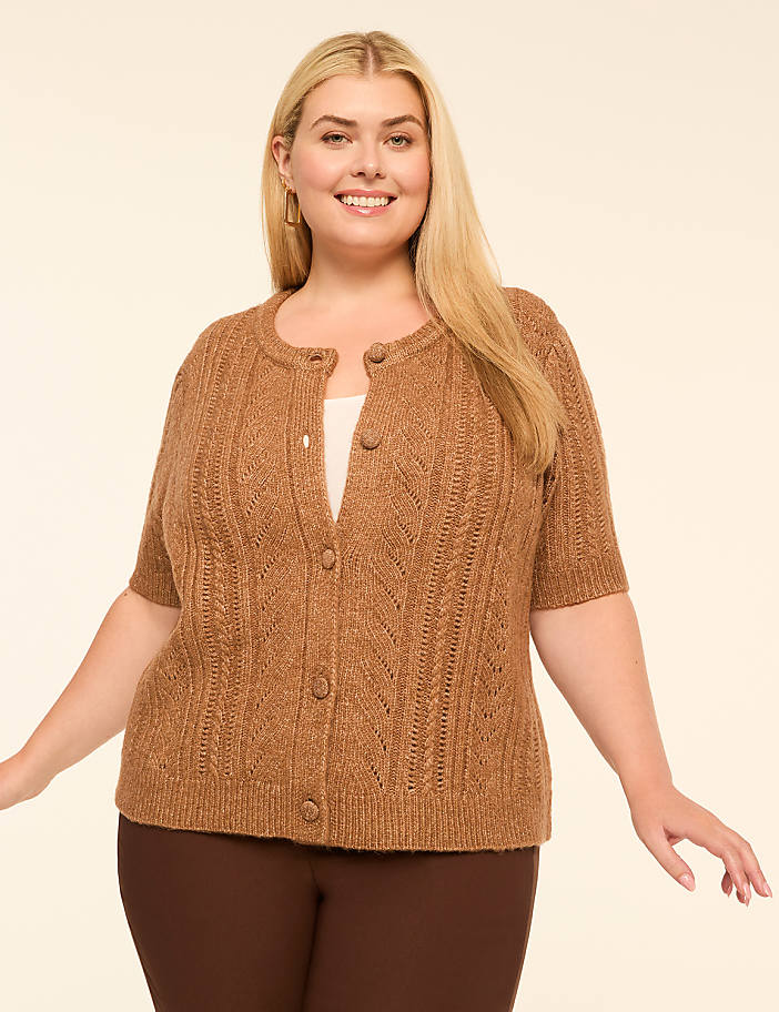 A smiling woman in a brown cardigan stands against a plain, light-colored background.