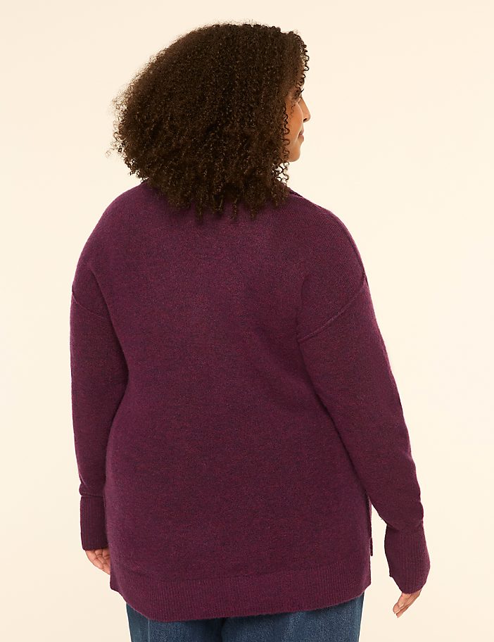A woman with curly brown hair, seen from the back, wears a plum-colored sweater and blue jeans against a plain, light-colored background.