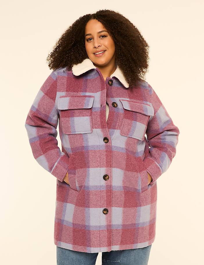 A smiling woman with curly hair stands in the foreground wearing a plaid jacket with a cream collar, against a plain cream background.