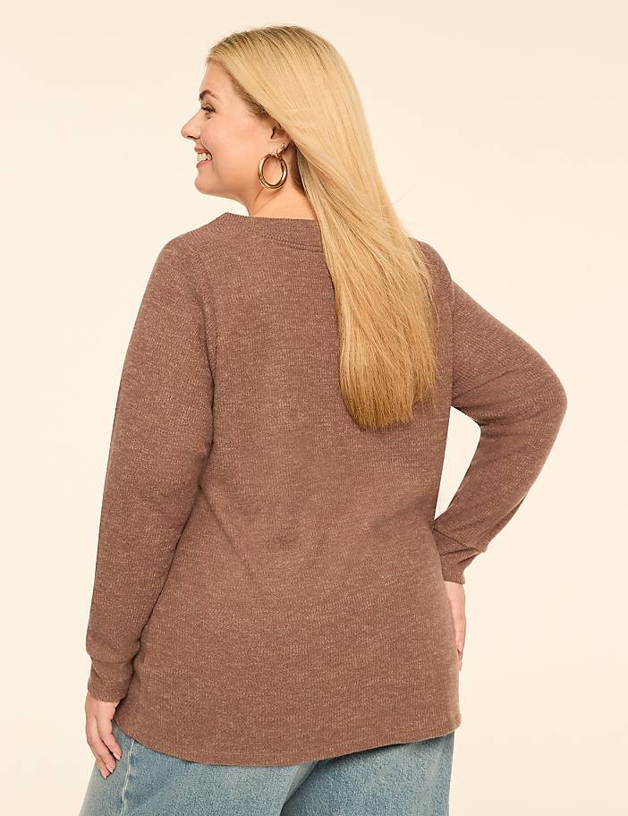 A woman with blonde hair wearing a brown sweater and blue jeans stands against a plain cream background.