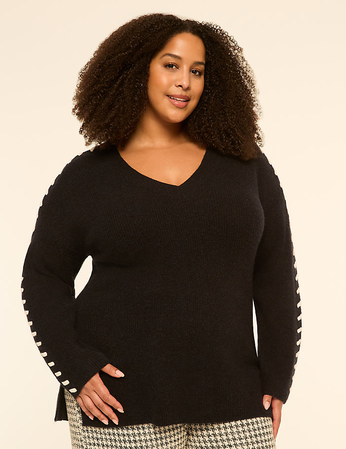 A woman with curly hair and a v-neck sweater stands against a plain, light-colored background.