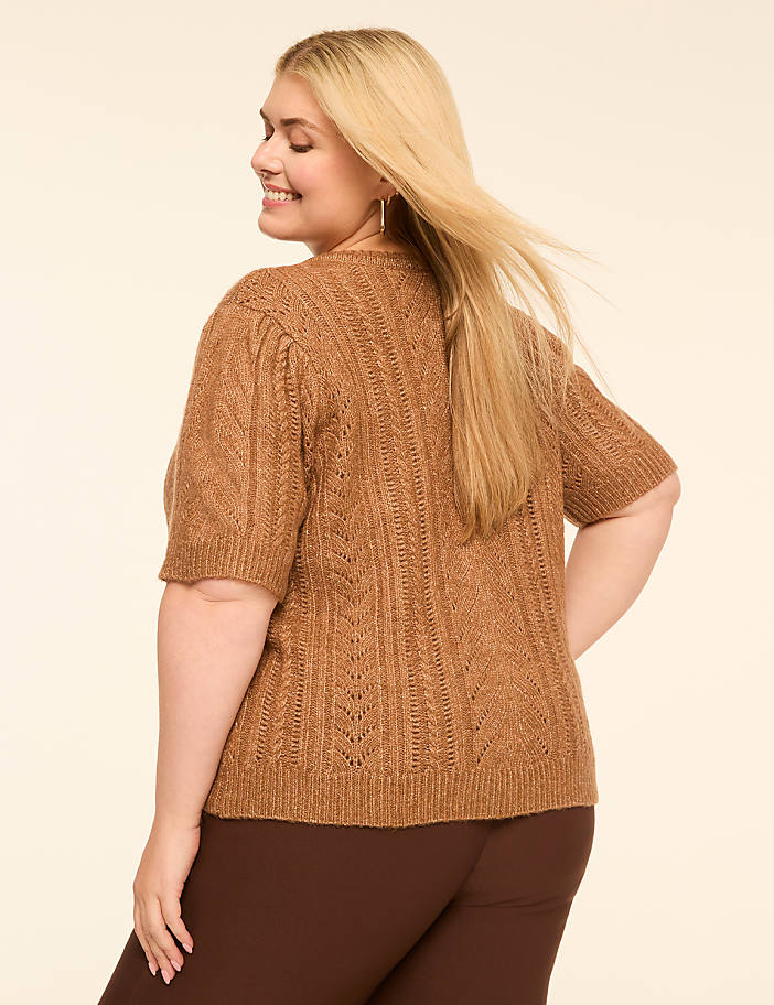A woman in a brown knit sweater and dark pants smiles over her shoulder against a plain, light background.